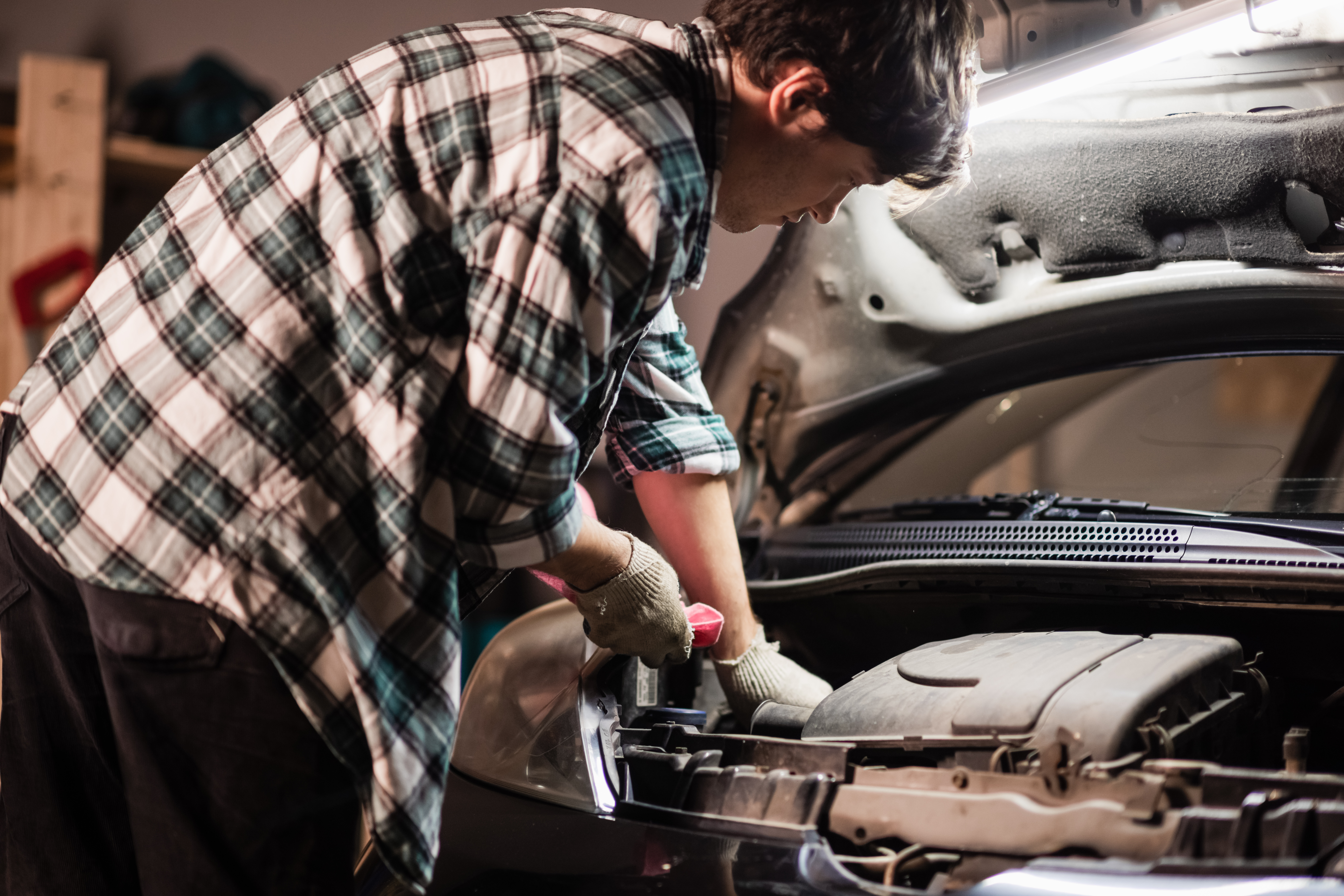 a young male car mechanic in a shirt and gloves checks the engine oil level in a black car.